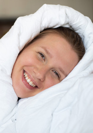 happy lazy sleepy girl, young positive cheerful woman in bed in bedroom covering herself in blanket, enjoying early morning, day at home in living room. Staying at home at weekend. Vertical photoの写真素材