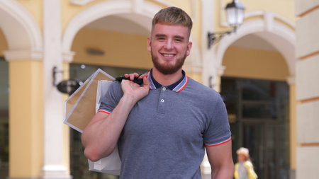 Portrait of happy positive stylish bearded man, young guy smiling in mall shopping, carrying, holding shopping paper bags in hand outdoors at summer sunny dayの写真素材