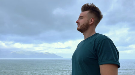 Profile side photo of handsome happy guy, young man enjoying sea, ocean view, landscape, looking into distance at summer day in tropical country at storm with cloudy sky, breathing deep fresh airの写真素材