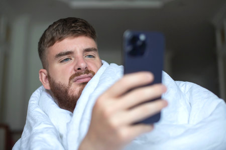 young unhappy angry annoyed man covering himself in blanket with sad frustrated look at home in living room in early morning talking on cell mobile phone smartphone, having negative conversation callの写真素材