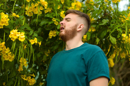 Portrait of handsome young allergic man is suffering from pollen allergy or cold on natural flowers,...