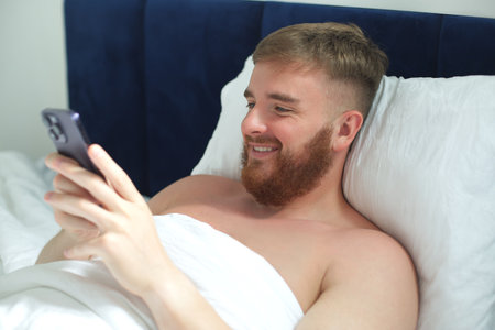 Young handsome man in the bed in bedroom at home in the morning lying under white blanket using cell...