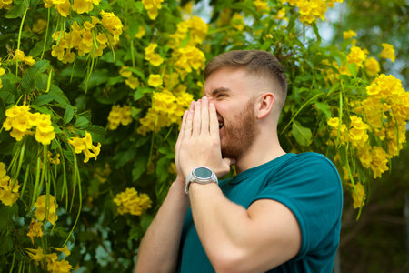 Portrait of handsome young allergic man is suffering from pollen allergy or cold on natural flowers, flowering tree background at spring or sunny summer day, sneezes, blowing his runny nose rubs eyesの写真素材