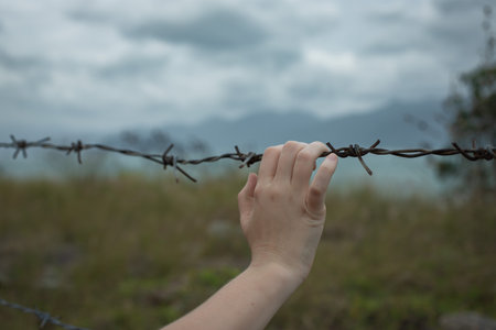a hand holds on to barbed wire against a stormy sky. man seeks freedom behind the fence.の写真素材
