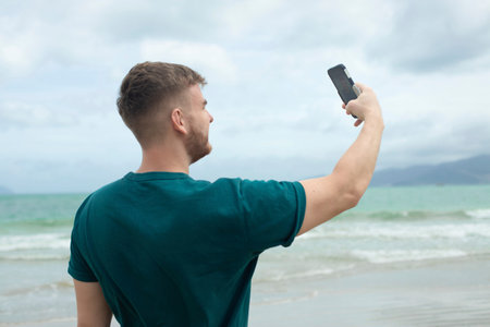 Young guy, handsome happy man at sea is taking picture of hisself, selfie at camera of his phone, using smartphone for social media at summer beach in tropical exotic country. Blogger, vlogの写真素材