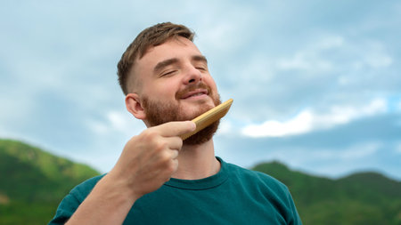 Handsome young bearded guy, European brutal man with a beard hair is combing the beard on face with a comb, brush. Male personal self care, products, accessory on nature. Natural summer backgroundの写真素材