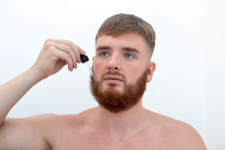 young handsome bearded man apply serum oil, cosmetic product, lotion for perfect fresh skin hydration, grow beard, using skincare, hydrating on white backgroundの写真素材