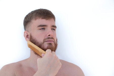 Handsome young bearded guy, European brutal man with a beard hair is combing the beard on face with a comb, brush. Male personal self care, products, accessory isolated on white backgroundの写真素材