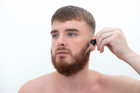 young handsome bearded man apply serum oil, cosmetic product, lotion for healthy perfect fresh skin hydration, grow beard, using skincare, hydrating on white backgroundの写真素材