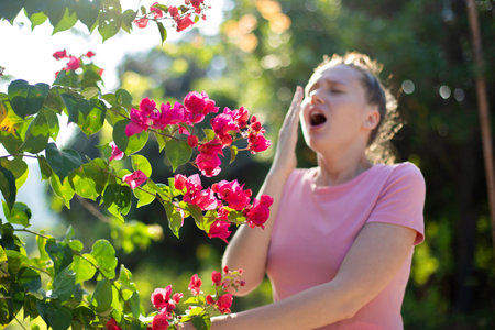 Portrait of beautiful young allergic woman is suffering from pollen allergy or cold on natural flower flowering tree background at spring or sunny summer day sneezes, blowing her runny nose rubs eyesの写真素材