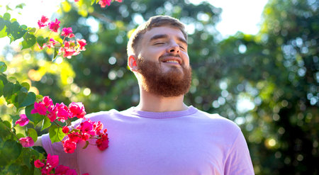 Portrait of happy handsome bearded guy, young positive man with beard is smelling beautiful red pink flowers in the garden, smiling, enjoying spring or summer day, breathing deep deeply fresh airの写真素材