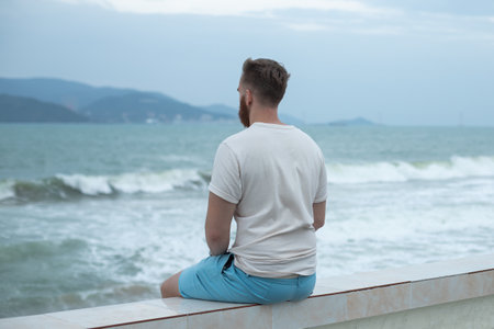 Young calm sad serious man is sitting on embankment near sea, ocean on beach, thinking, meditate on natural backgroundの写真素材