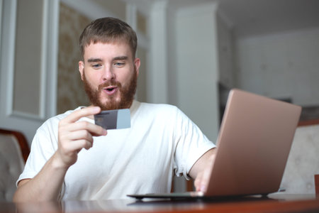 Happy young man hold credit bank card in hand, use laptop computer, smile at home, shopping online, purchase in internetの写真素材