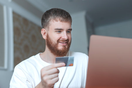 Happy young man hold credit bank card in hand, use laptop computer, smile at home, shopping online,...