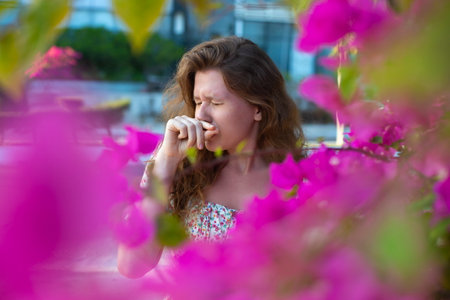 Portrait of beautiful young allergic woman is suffering from pollen allergy or cold on natural flower flowering tree background at spring or sunny summer day sneezes, blowing her runny nose rubs eyesの写真素材