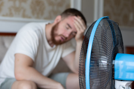 Young bearded man using electric fan at home, sitting on couch cooling off during hot weather, suffering from heat, high temperatureの写真素材