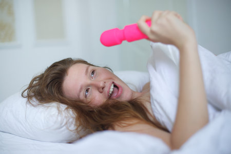 Young happy sexy woman hold in hand sex toy, masturbate with vibrator in bed in bedroom at home.の写真素材