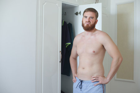 Unhappy sad upset young man with fat on her stomach, guy worried about body fat touching belly standing at home near closetの写真素材