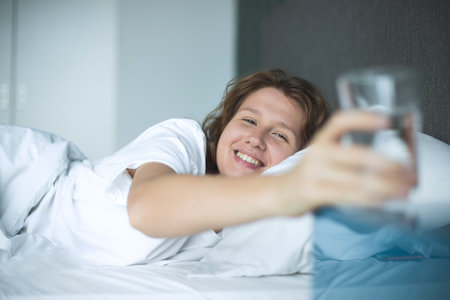 Young woman in the bed in bedroom at home in the morning lying under white blanket and drinking pure fresh water from glassの写真素材