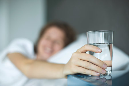 Young woman in the bed in bedroom at home in the morning lying under white blanket and drinking pure fresh water from glassの写真素材