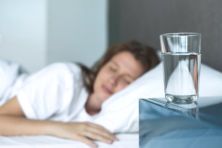 Young woman in the bed in bedroom at home in the morning lying under white blanket and drinking pure fresh water from glassの写真素材