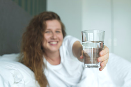 Young woman in the bed in bedroom at home in the morning lying under white blanket and drinking pure fresh water from glassの写真素材