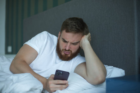 Young bearded man is using cell mobile phone at home in bed in bedroom, looking at screen of...