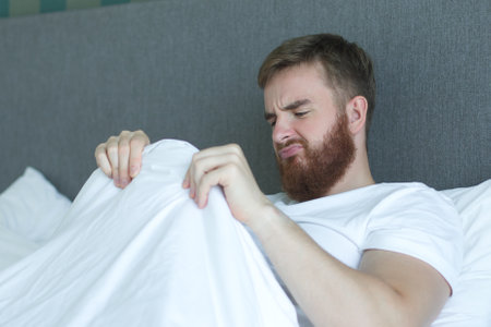 Young man worrying about his penis and erection failure, surprised guy is having trouble with his potency in bed in bedroomの写真素材