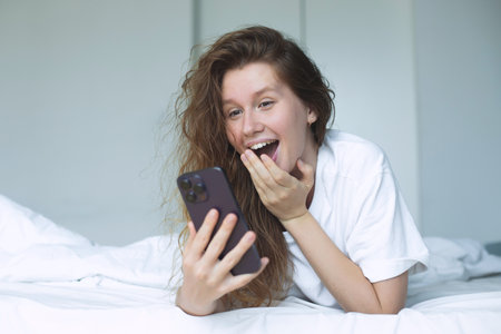 young happy woman use phone and lying in bed in bedroom in the morning, concept morning, phone addiction, social, habitsの写真素材