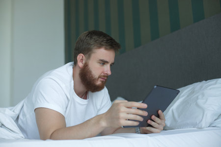 Young happy man use tablet laptop computer at home in bed, lay in bedroom, looking at screen of...