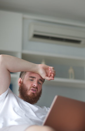 Young man with air conditioner remote control using air conditioner at home, guy is cooling off during hot weather at summer, suffering from heat, high temperature. Broken ACの写真素材