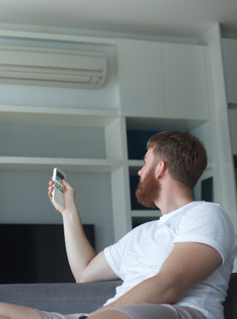 Young man with air conditioner remote control using air conditioner at home, guy is cooling off during hot weather at summer, suffering from heat, high temperature. Broken ACの写真素材
