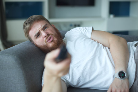portrait of joyful young man in casual clothes sitting and laying on sofa in living room and looking...