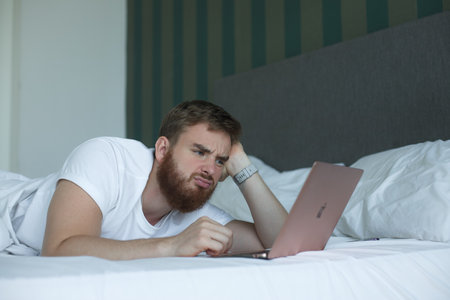 03/01/23 - Vietnam, Nha Trang: Young sad unhappy man use MacBook Apple laptop computer, gadget tablet at home in bed, lay in bedroom, looking at screen. Dissatisfied customer doesn't like the productのeditorial素材