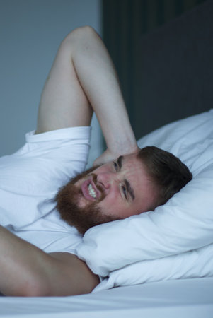 Young sad depressed unhappy worried man in the bed in bedroom at home in the morning lying under white blanket suffering from insomnia problems with sleep at nightの写真素材