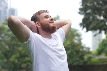 Positive young man smiling during the rain in the park. Cheerful male enjoying the rain outdoors....