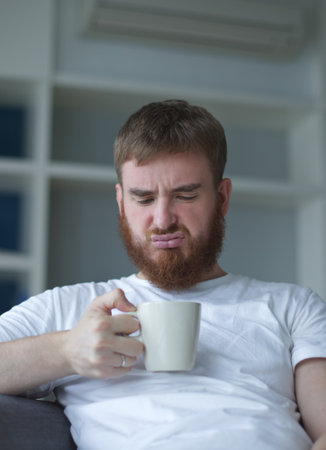 Unsatisfied, sad, upset man with mug, man does not want to take the medicine, beverage man is sitting in the living room, at homeの写真素材