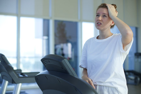 Tired exhausted young woman is training workout, running on treadmill in gym holding her head with...