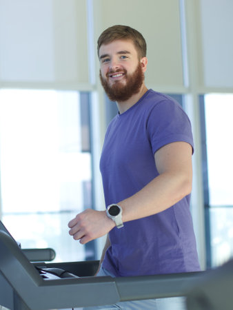 Happy young fit man training in the gym, running jogging on treadmill, doing cardio exercise and smile. Healthy lifestyle, sport, fitness concept, vertical photoの写真素材