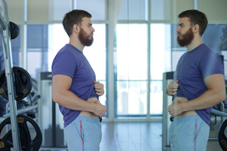 Young sad upset man is disappointed because of fat belly, stomach, looking at mirror with frustrated look, trying to loose weightの写真素材