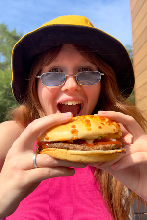 Portrait of young happy woman is eating, biting juicy delicious burger outside cafe looking at camera and smile. Fast junk food, unhealthy eating concept.の写真素材