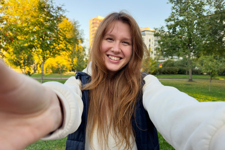 Portrait of happy cheerful young beautiful woman walking outdoors at summer or golden autumn park, smile and look at camera, hug or take selfie on cameraの写真素材