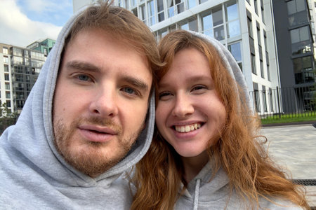 Smiling young couple looking at screen of mobile phone while and taking selfie against city buildings, concept love, young familyの写真素材