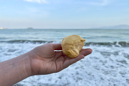 a mans hand holds a piece of bad-smelling ripe durian against the background of the sea, ocean,...