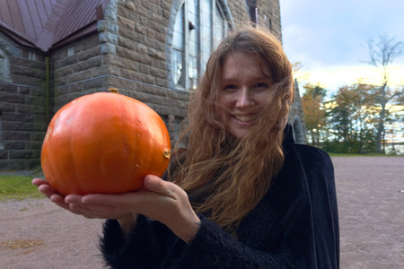 Young woman witch in a black robe with Halloween pumpkin on the church backgroundの写真素材