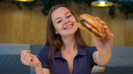 Portrait of young happy woman or beautiful teenager girl eating fast junk food, tasty burger and drink soda in a restaurant or cafe, enjoy meal.の写真素材