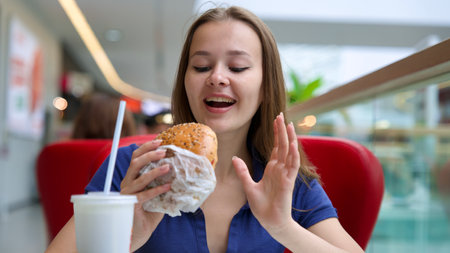 Portrait of young happy woman or beautiful teenager girl eating fast junk food, tasty burger and drink soda in a restaurant or cafe, enjoy meal.の写真素材