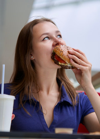 Portrait of young happy woman or beautiful teenager girl eating fast junk food, tasty burger and drink soda in a restaurant or cafe, enjoy meal.の写真素材