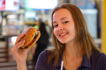 Portrait of young happy woman or beautiful teenager girl eating fast junk food, tasty burger and drink soda in a restaurant or cafe, enjoy meal.の写真素材