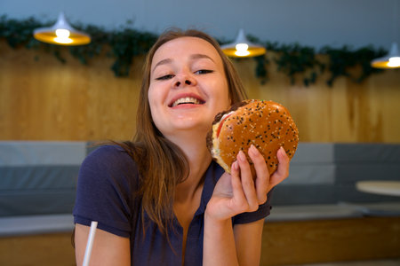 Portrait of young happy woman or beautiful teenager girl eating fast junk food, tasty burger and drink soda in a restaurant or cafe, enjoy meal.の写真素材